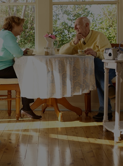 Una pareja de ancianos habla sentada en una mesa. El dispositivo de terapia ResMed se encuentra sobre una mesa en la esquina