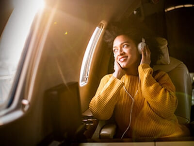 Una joven mirando por la ventanilla de un avión mientras lleva auriculares, con la luz del sol entrando por la ventanilla.